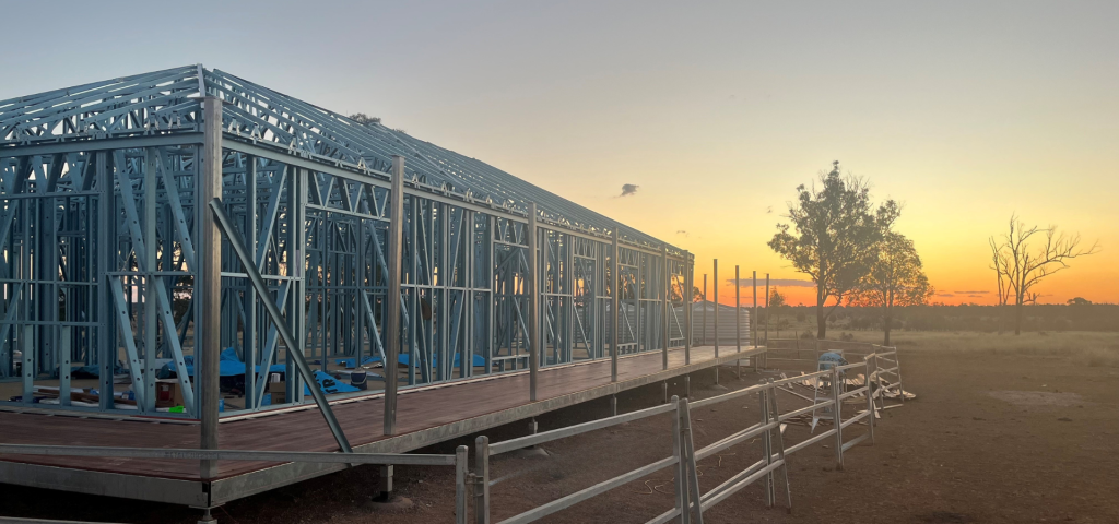Steel-framed building under construction on a raised deck at sunset; orange sky and sparse trees in the distance.